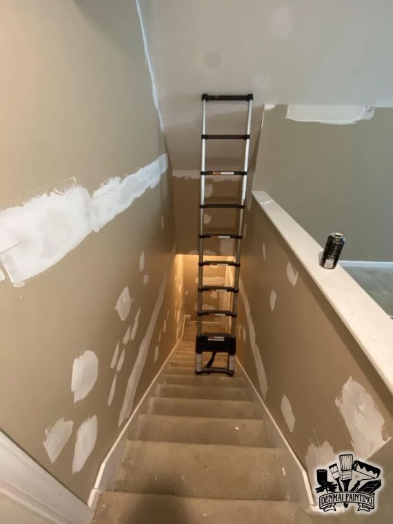 Stairwell under renovation with a ladder set over the steps and patchy drywall on beige walls