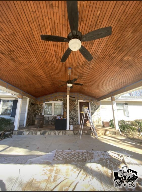 Covered patio with wood ceiling and ceiling fans, ladder, chair, and porch furniture in a sunlit yard.