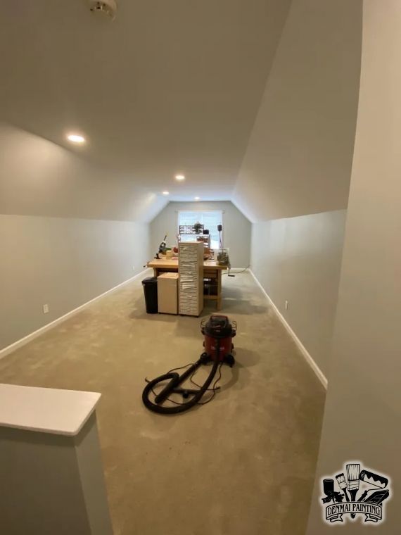 Unfinished attic room with beige carpet, white sloped walls, moving boxes, and a vacuum cleaner in the foreground