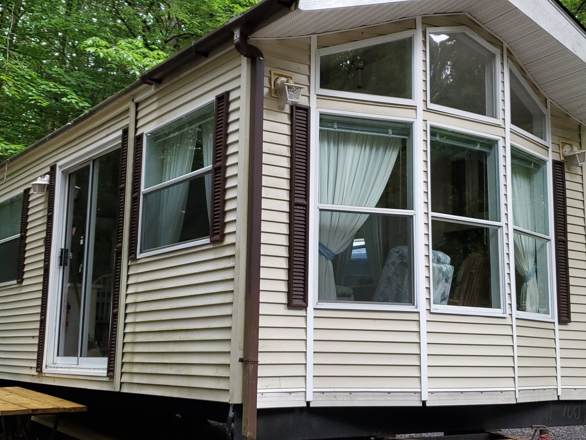 White-sided mobile home with a front bay window and porch in a wooded setting