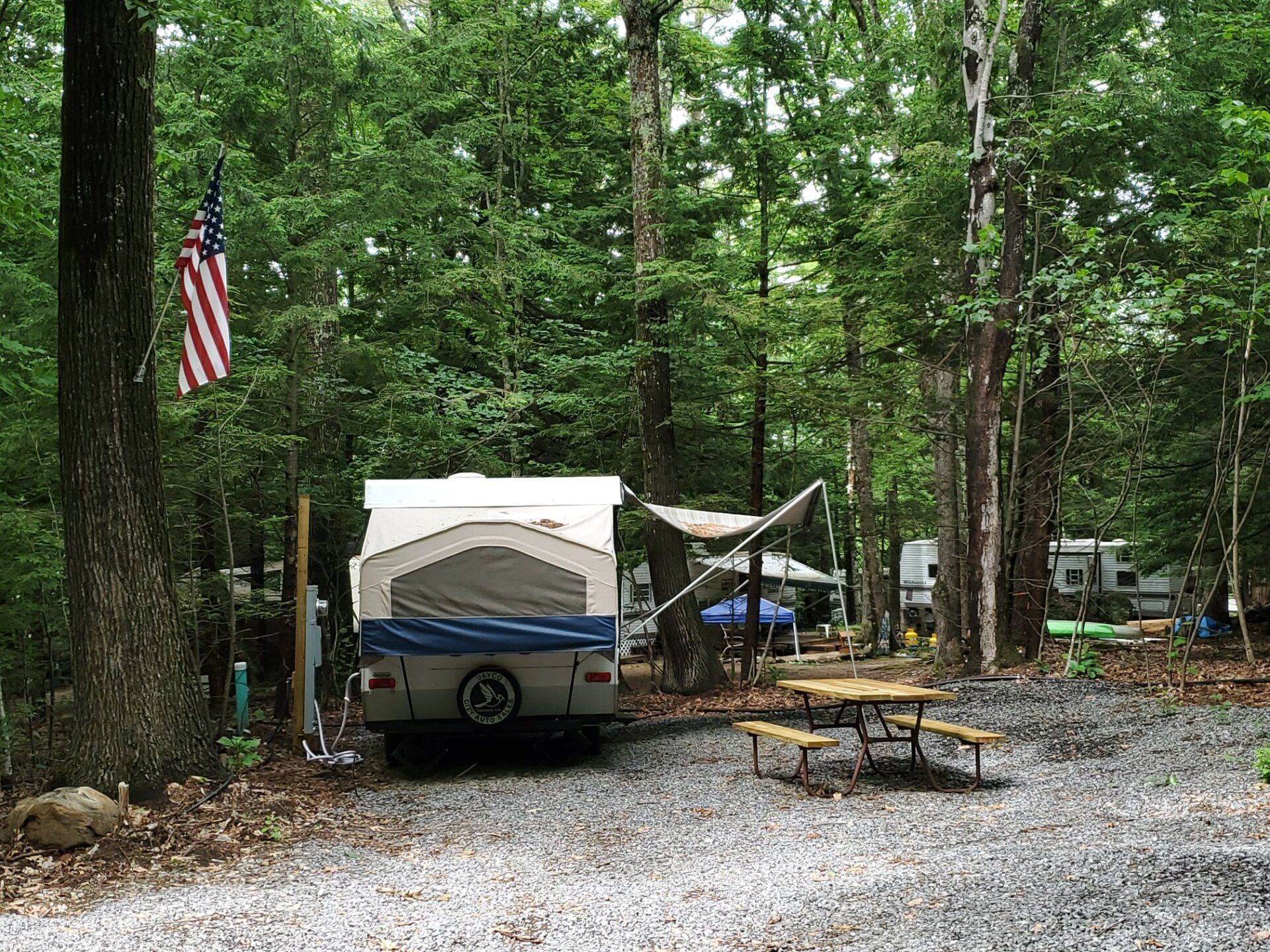 Campsite in wooded area with camper, American flag, picnic table, and a gravel fire ring area
