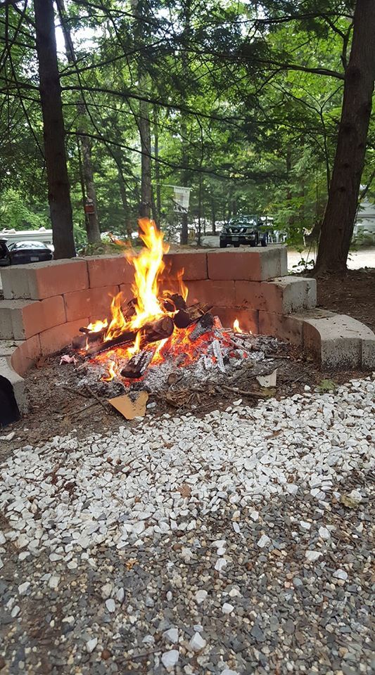 Small campfire burning in a brick fire pit on a gravel surface, with trees and parked cars in the background.