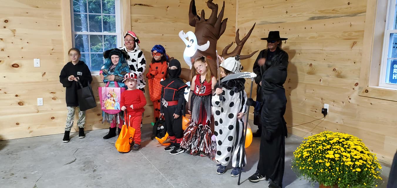 Group in colorful Halloween costumes posing indoors against a wooden wall, with pumpkins and flowers nearby