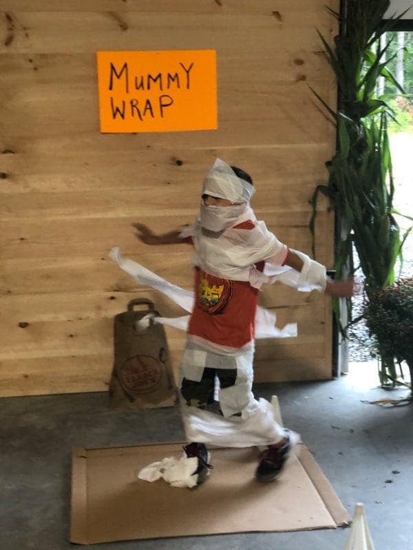 Person wrapped like a mummy standing in a box, with “MUMMY WRAP” sign on a wooden wall.