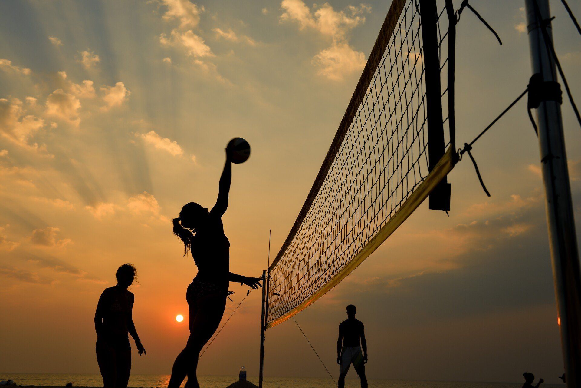 Silhouetted volleyball players at sunset, one jumping to spike the ball by the net