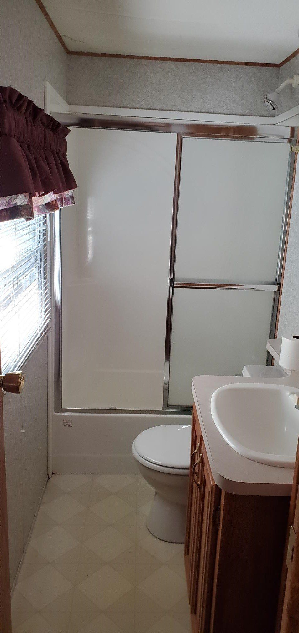 Small bathroom with shower curtain, toilet, sink, and window with maroon curtains.