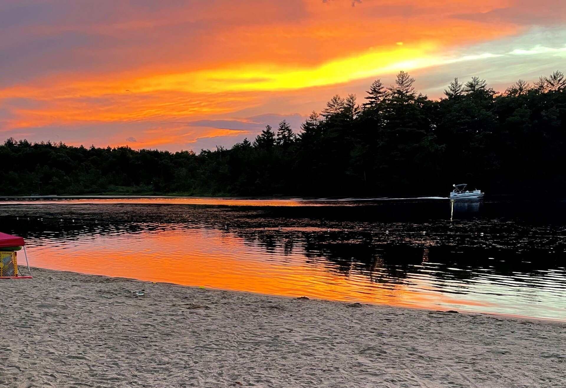 Sunset over a calm lake reflecting orange sky, with dark trees along the shore and a sandy beach in front