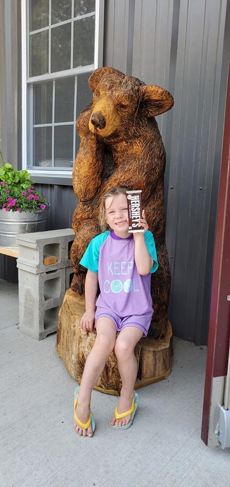 Child sitting on a wooden bear statue outside a building, holding a phone and smiling