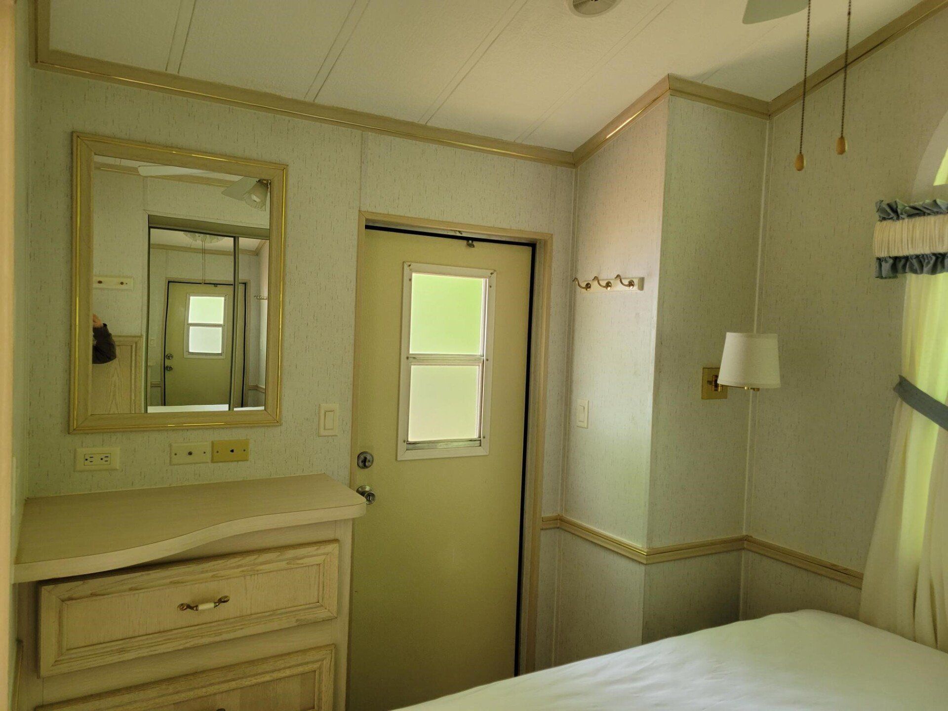 Small beige bedroom with dresser, mirror, door, and a bed by the window.