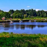 Calm pond with small boats, green grass, and trees under a clear blue sky