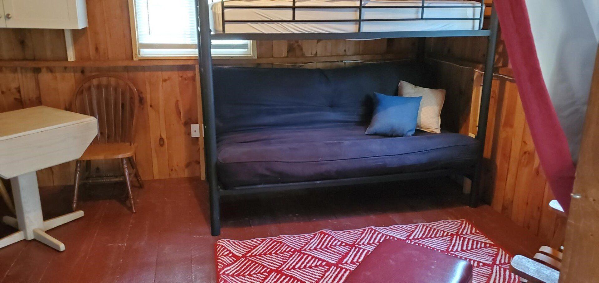 Wood-paneled room with a bunk bed, purple bedding, a small table, and a red patterned rug.