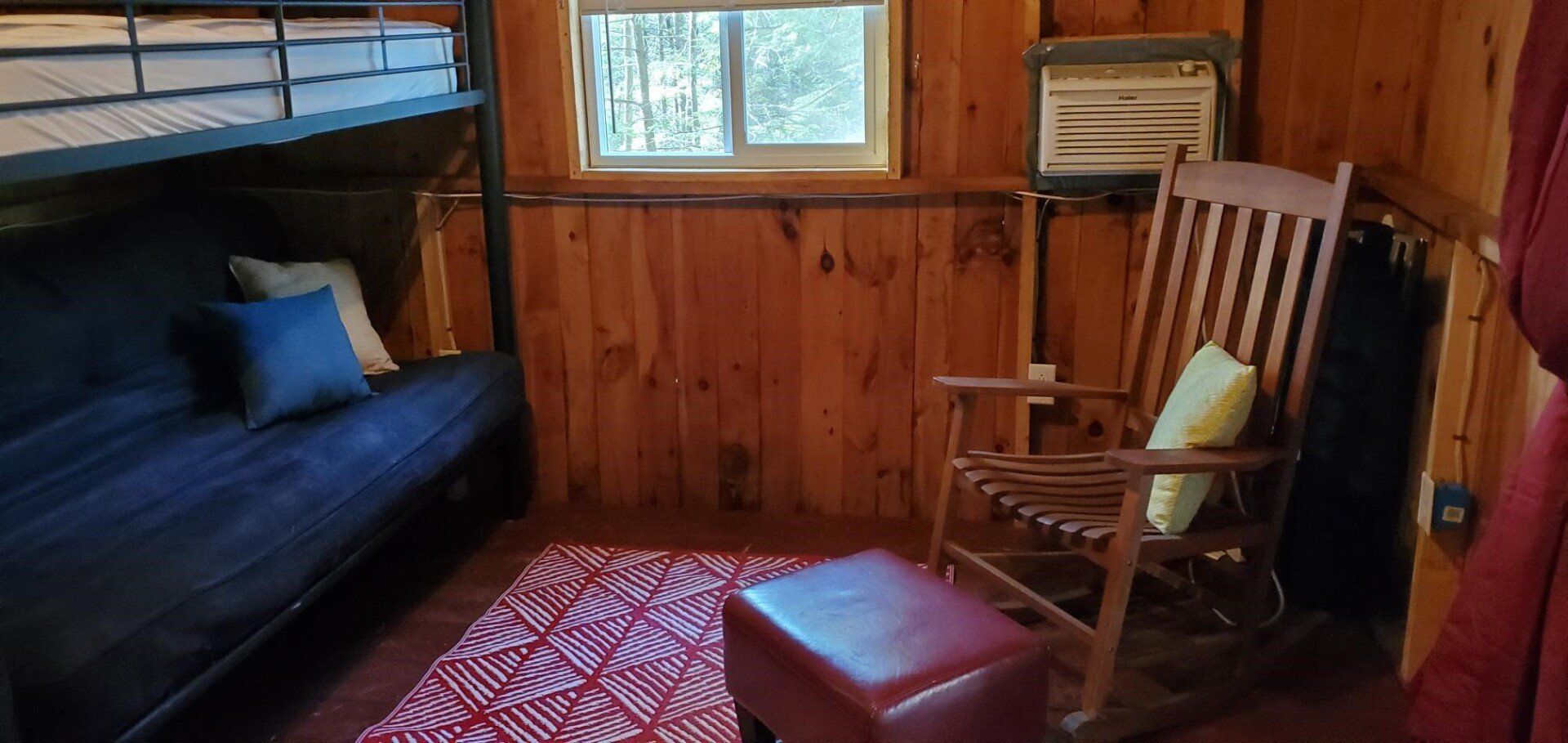 Cozy wood-paneled cabin room with bunk bed, rocking chair, small window, and red rug