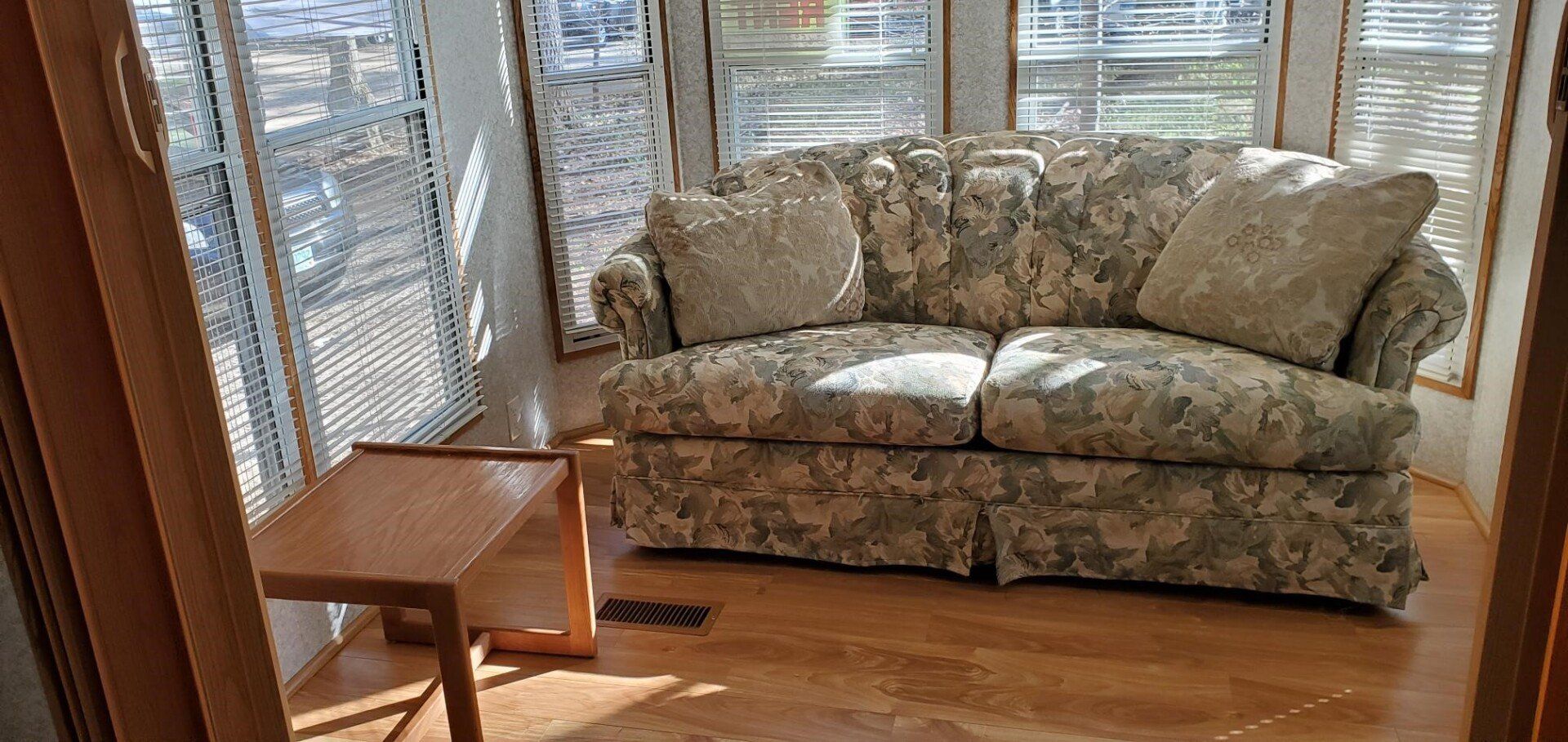 Sunlit living room with floral sofa, small wooden side table, and hardwood floor near blinds