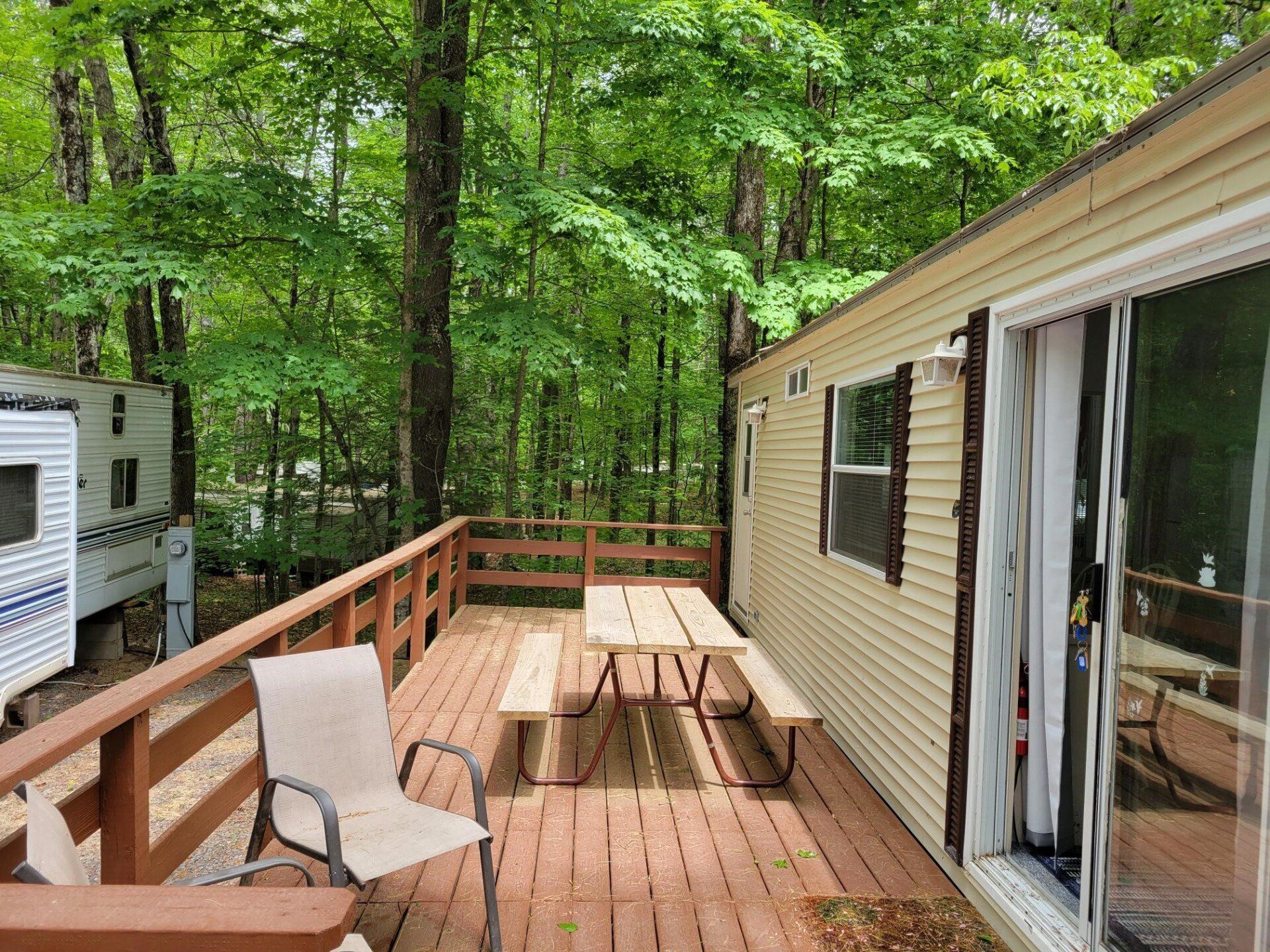 Wooden deck beside a beige cabin, with picnic table, chair, and wooded forest view.