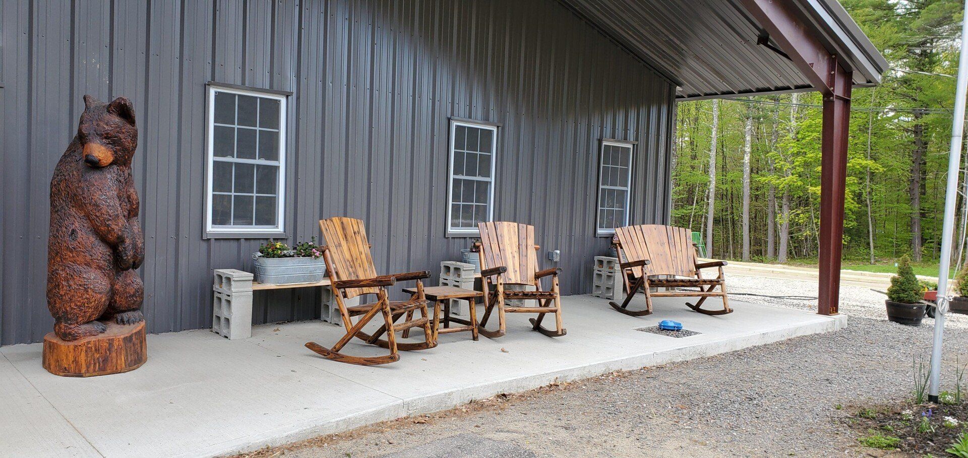 Covered porch with wooden rocking chairs beside a dark gray building and gravel driveway.