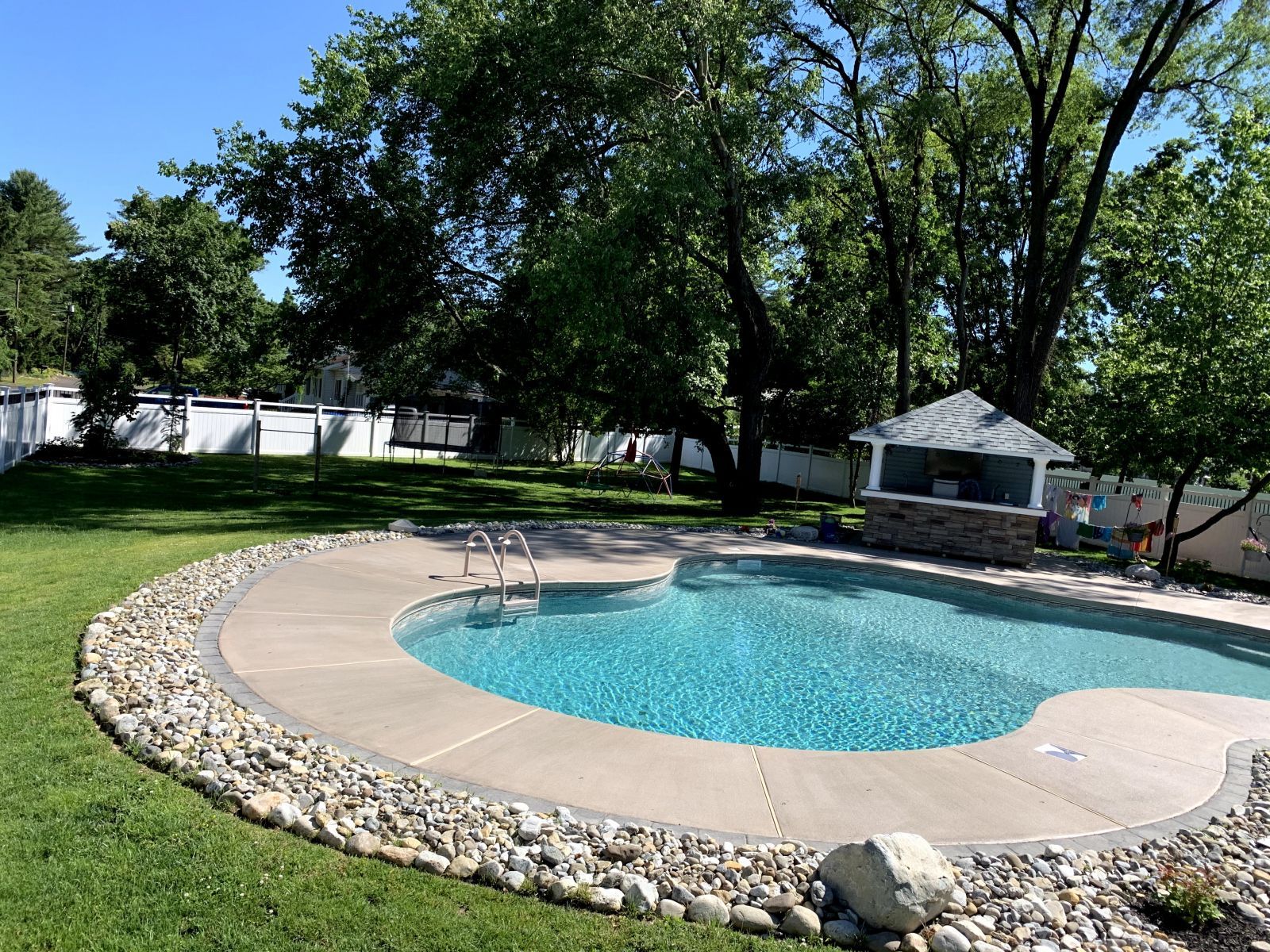 Backyard swimming pool with a curved stone border, green lawn, and trees on a sunny day