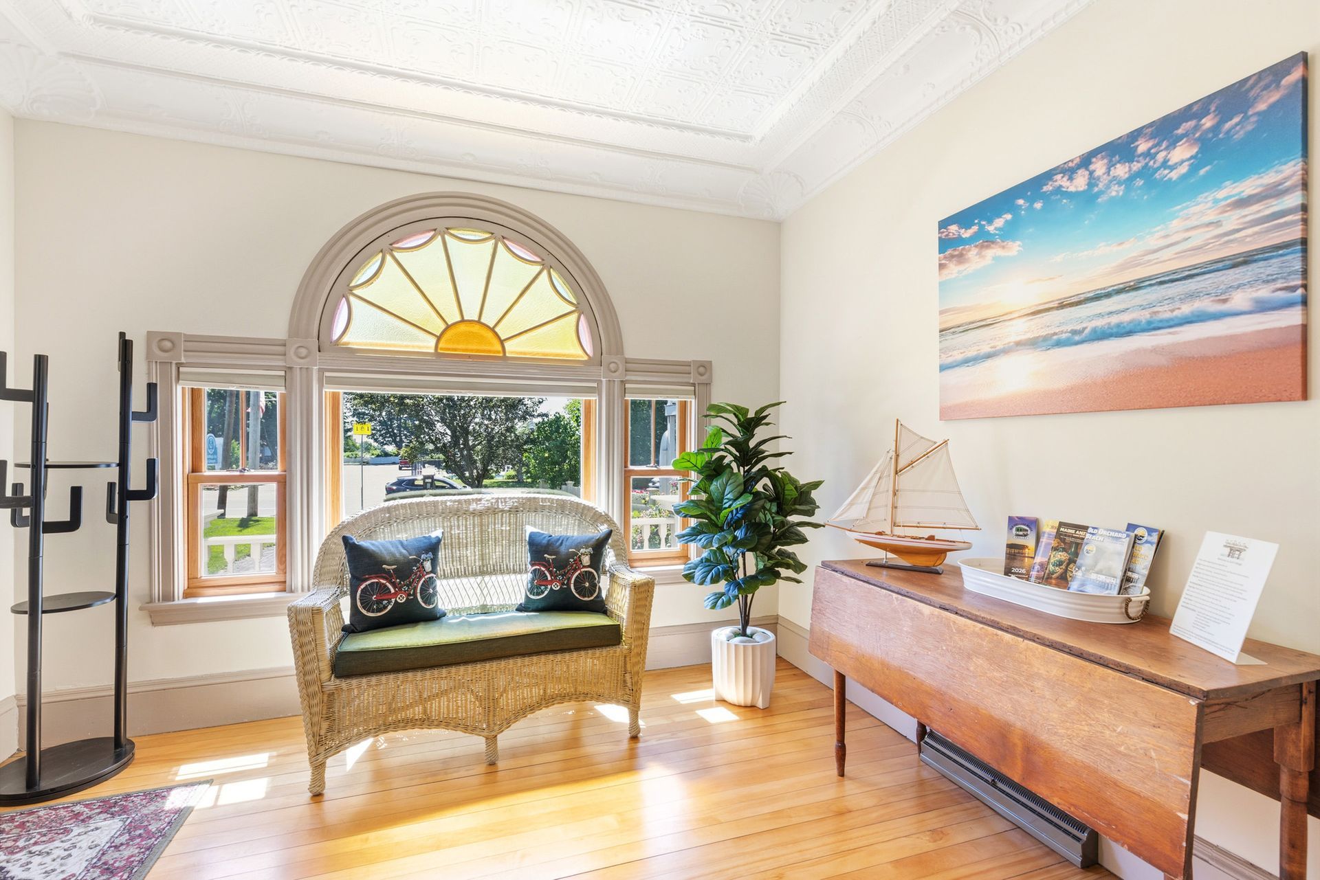 Bright sunroom with arched window, cushioned bench, wooden console table, beach painting, and sailboat decor.