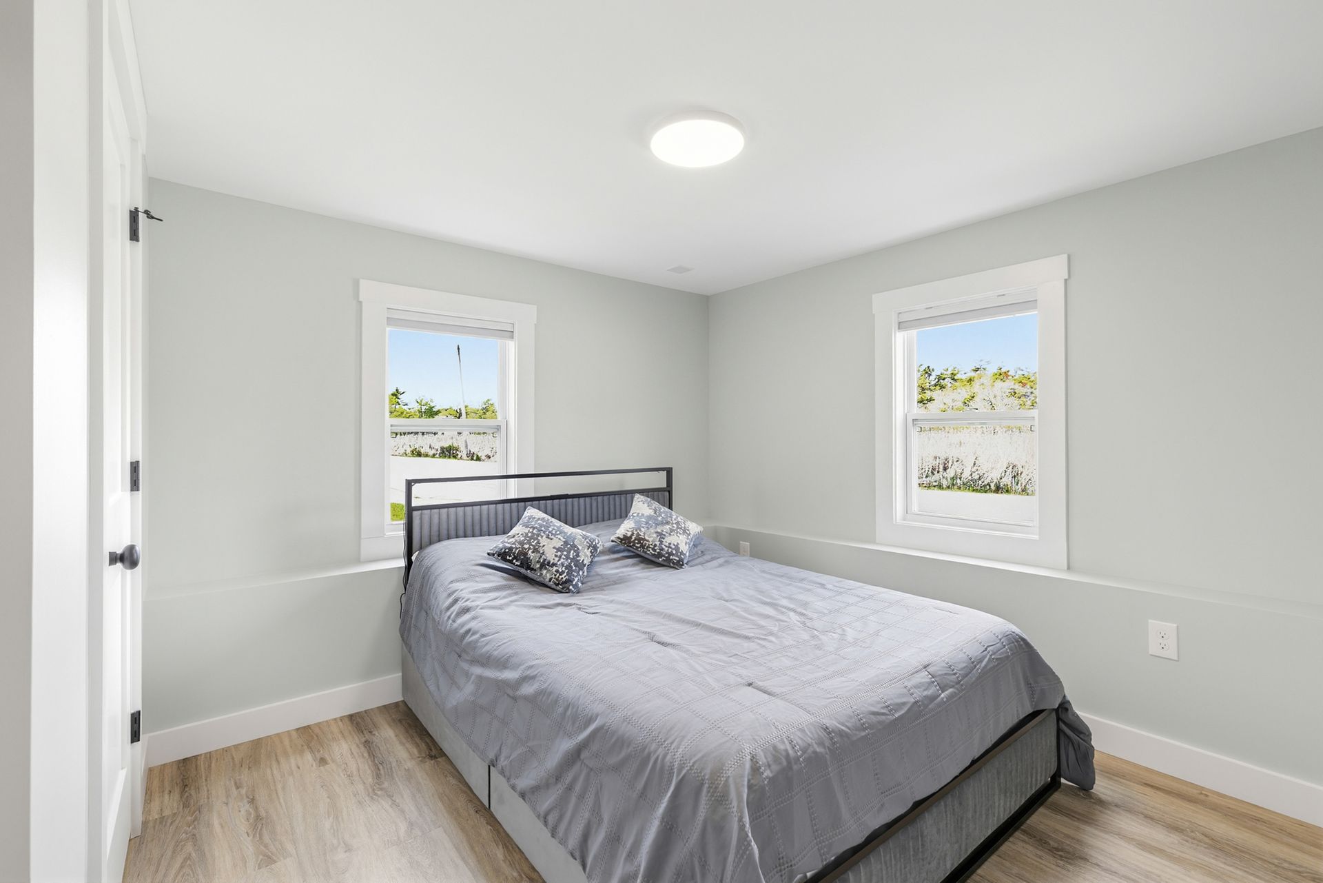Bright bedroom with a gray bed, white walls, two windows, and light wood floors