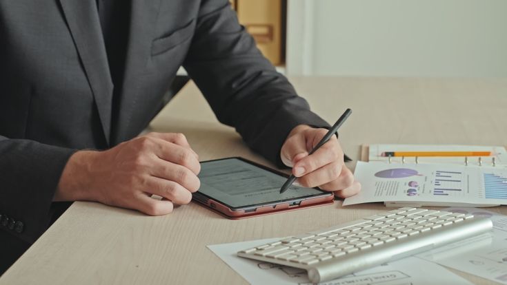 Person in suit using a stylus on a tablet at a desk with financial charts and keyboard.