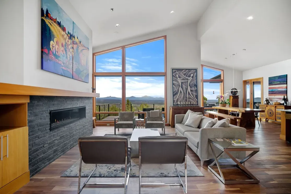 Spacious living room with large windows, fireplace, artwork, and a view of mountains under a blue sky.