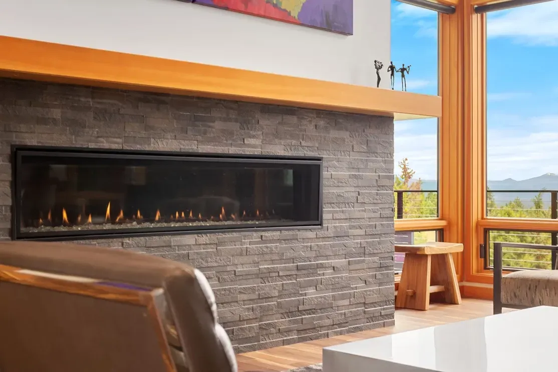 Modern fireplace with gray brick, long glass window, wooden accents, and a view of nature.