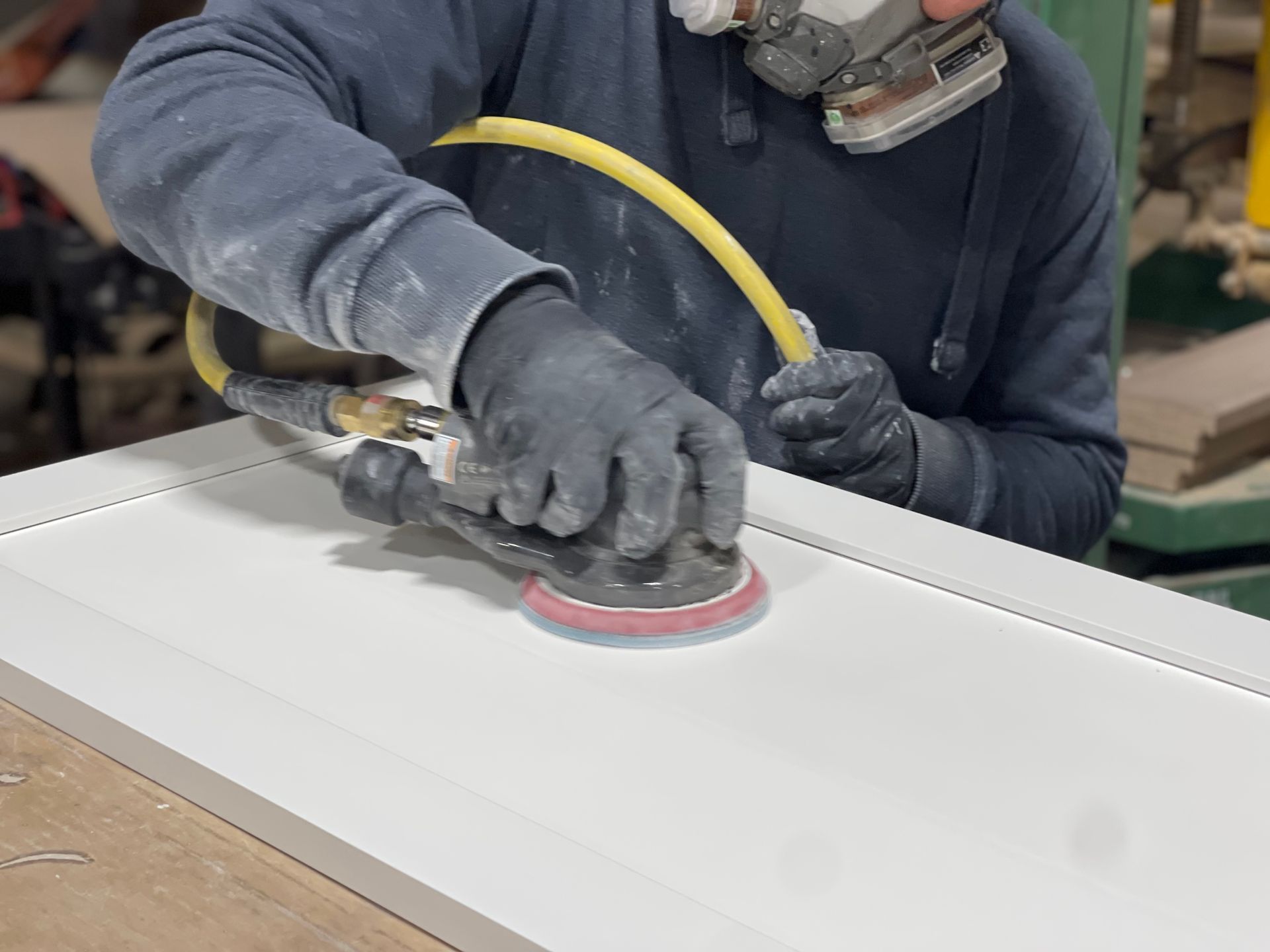 Person sanding a white surface with a power sander, wearing gloves and a respirator mask in a workshop.