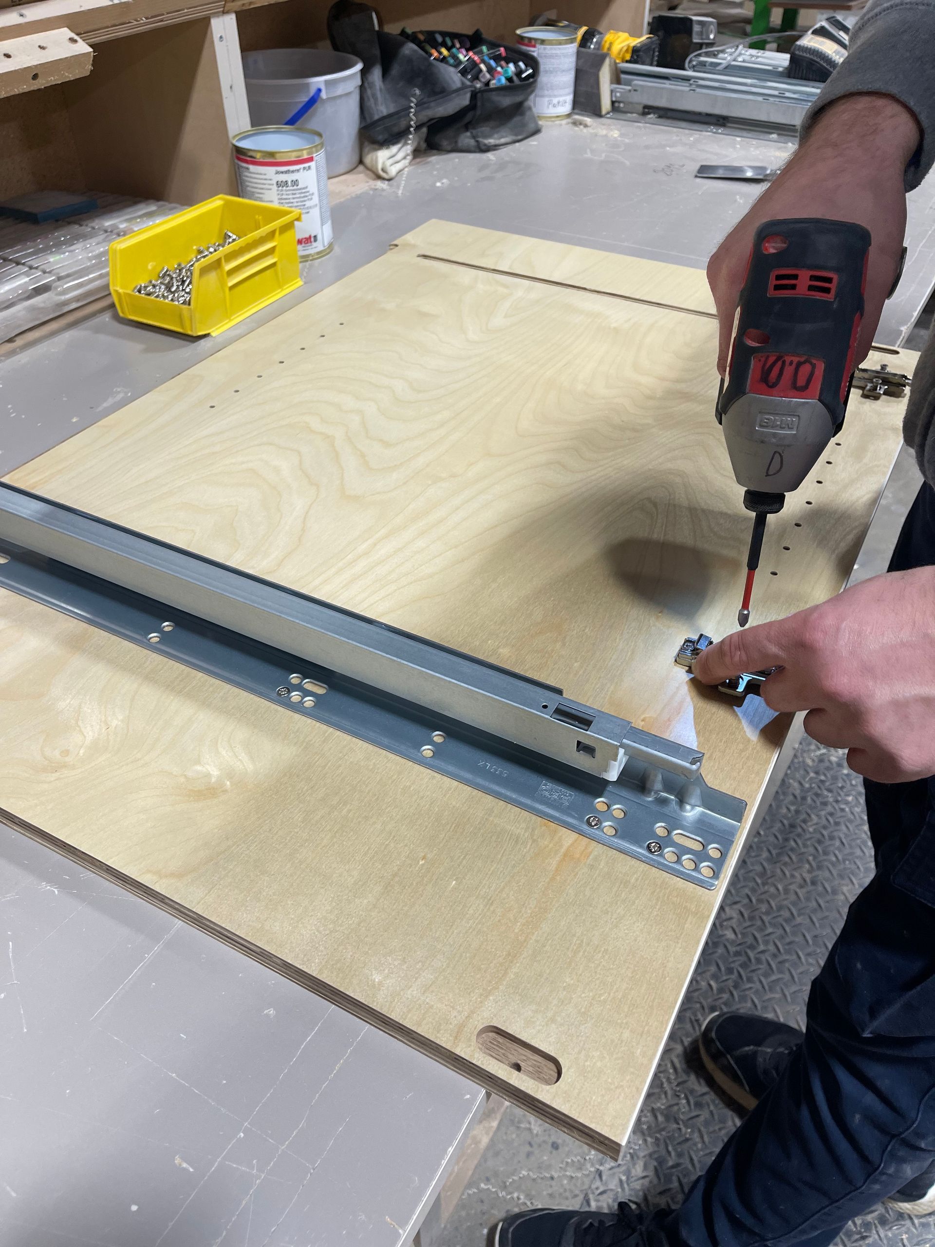 Person using a power drill to attach a metal drawer slide to a wooden panel in a workshop.