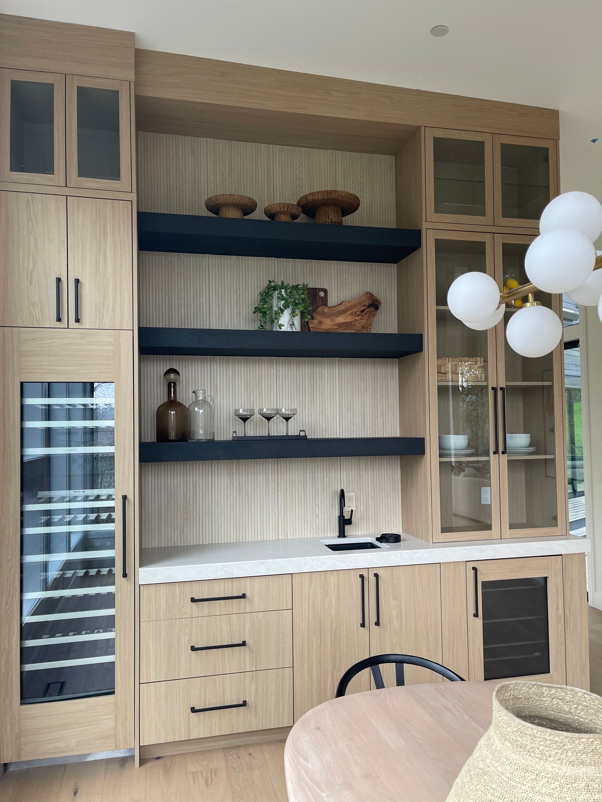 Modern light wood wet bar featuring black shelves, a wine cooler, glass-front cabinets, and a white countertop.