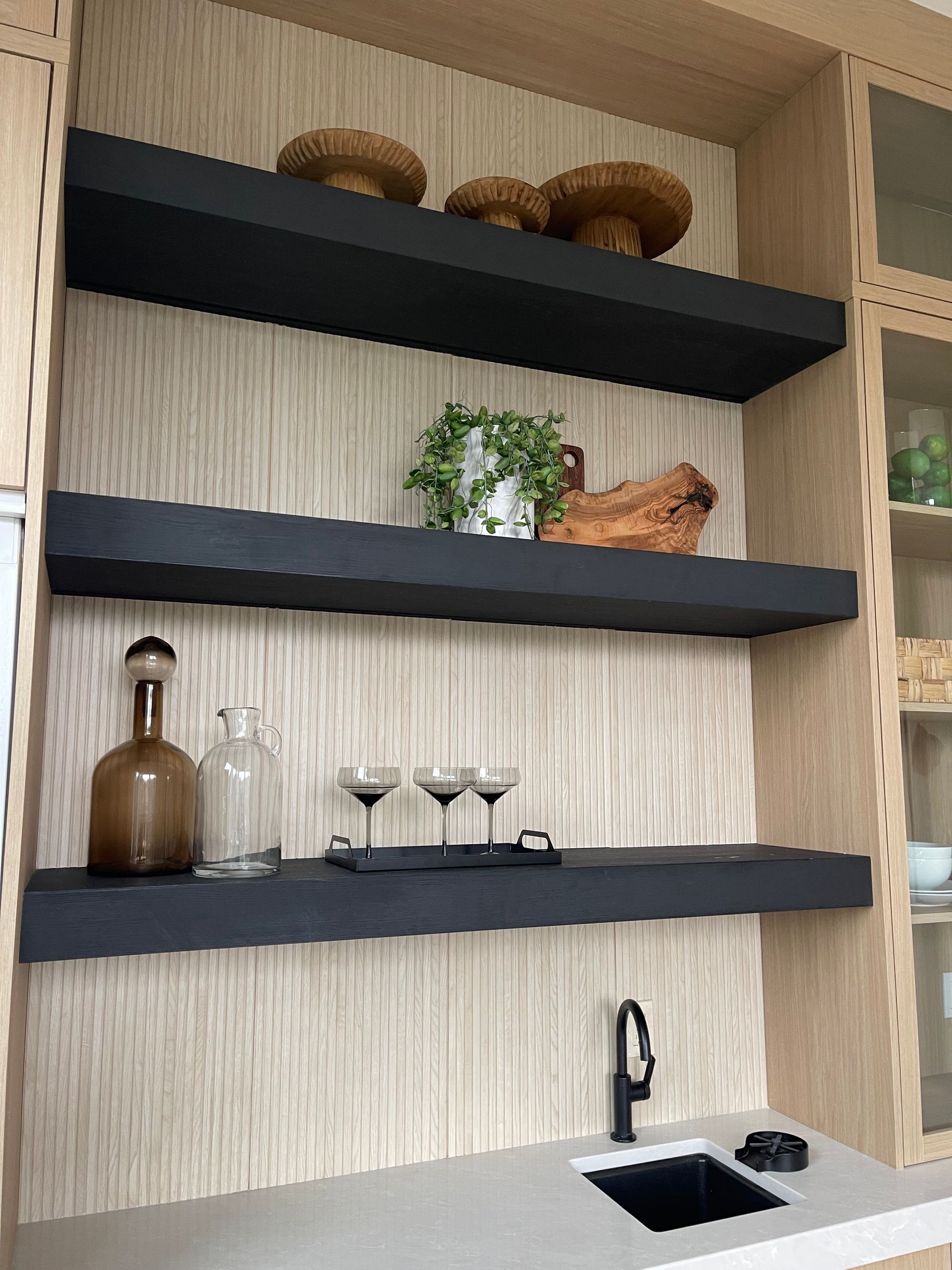 Three dark floating shelves against a light wood wall with decorative bowls, a plant, glassware, and a small kitchen sink.