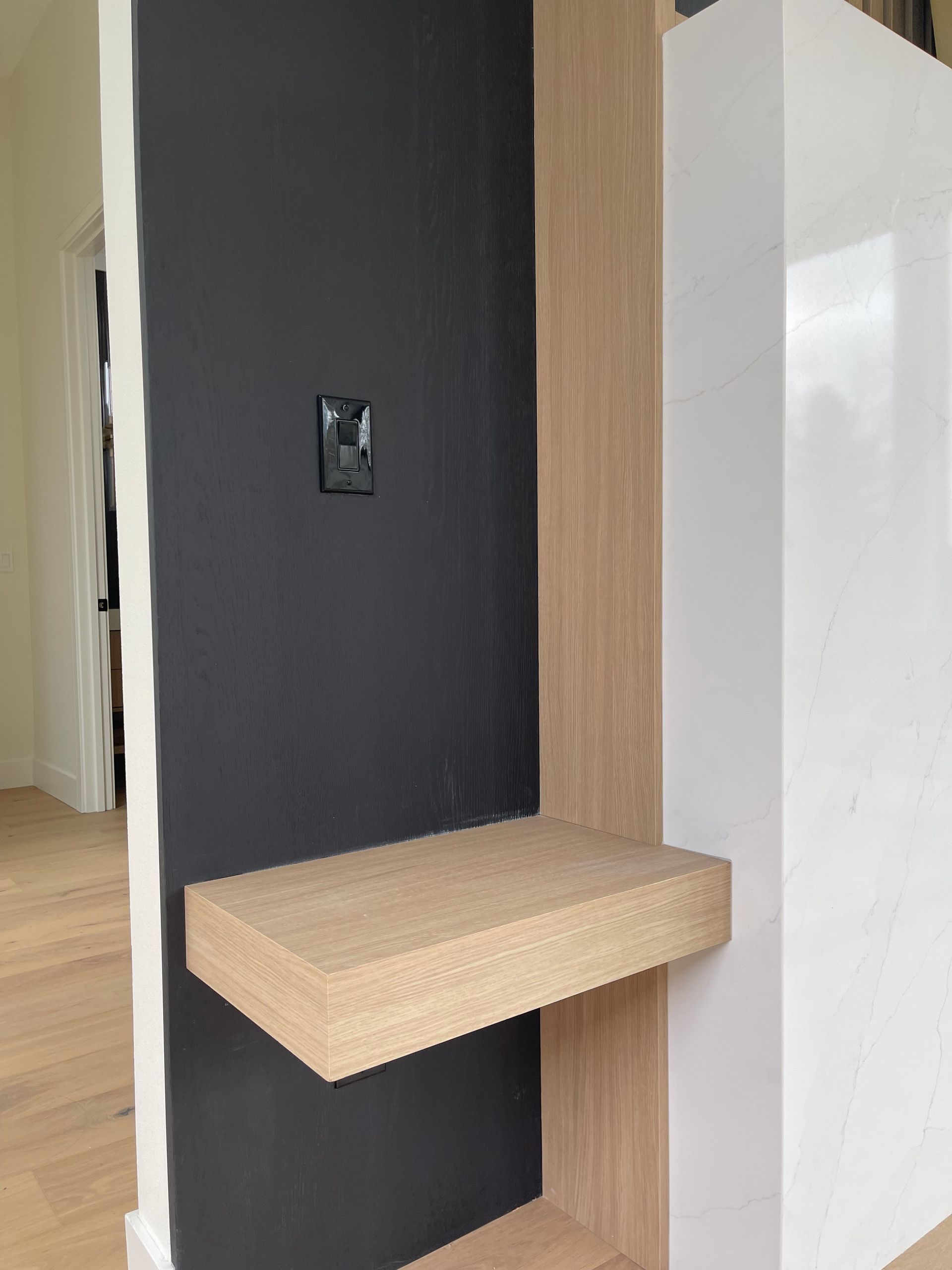 A floating wooden shelf mounted on a vertical wood panel against a dark grey wall next to a white surface.