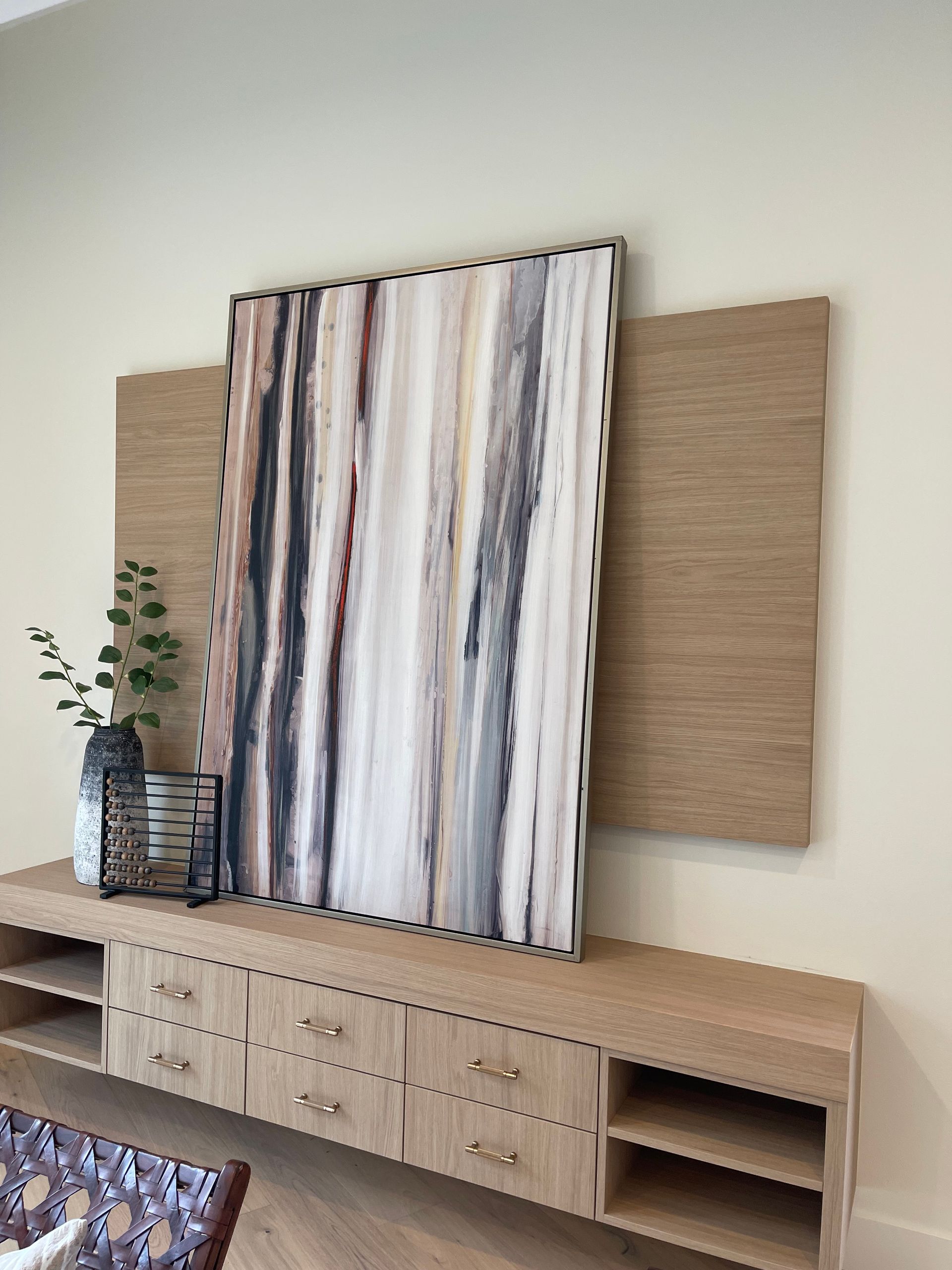 A vertical abstract art piece in a beige, brown, and grey palette rests on a wood media console against a wood wall panel.