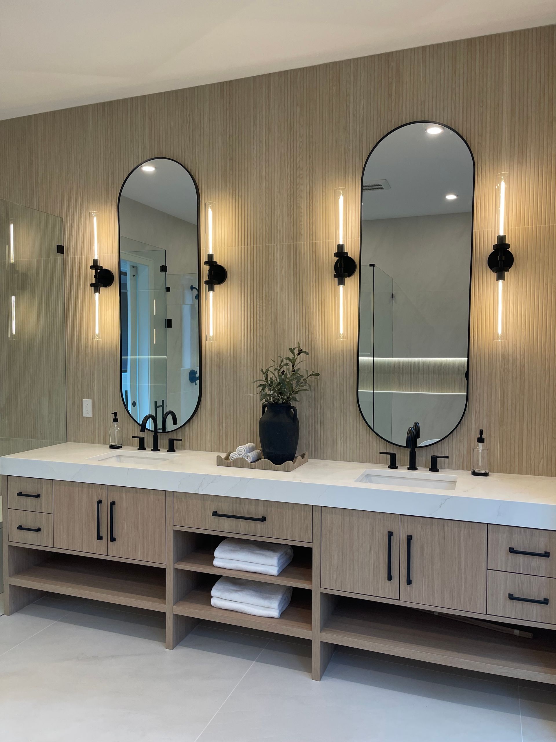 Double vanity bathroom with fluted wood walls, dual oval mirrors, modern sconce lighting, and white countertops.