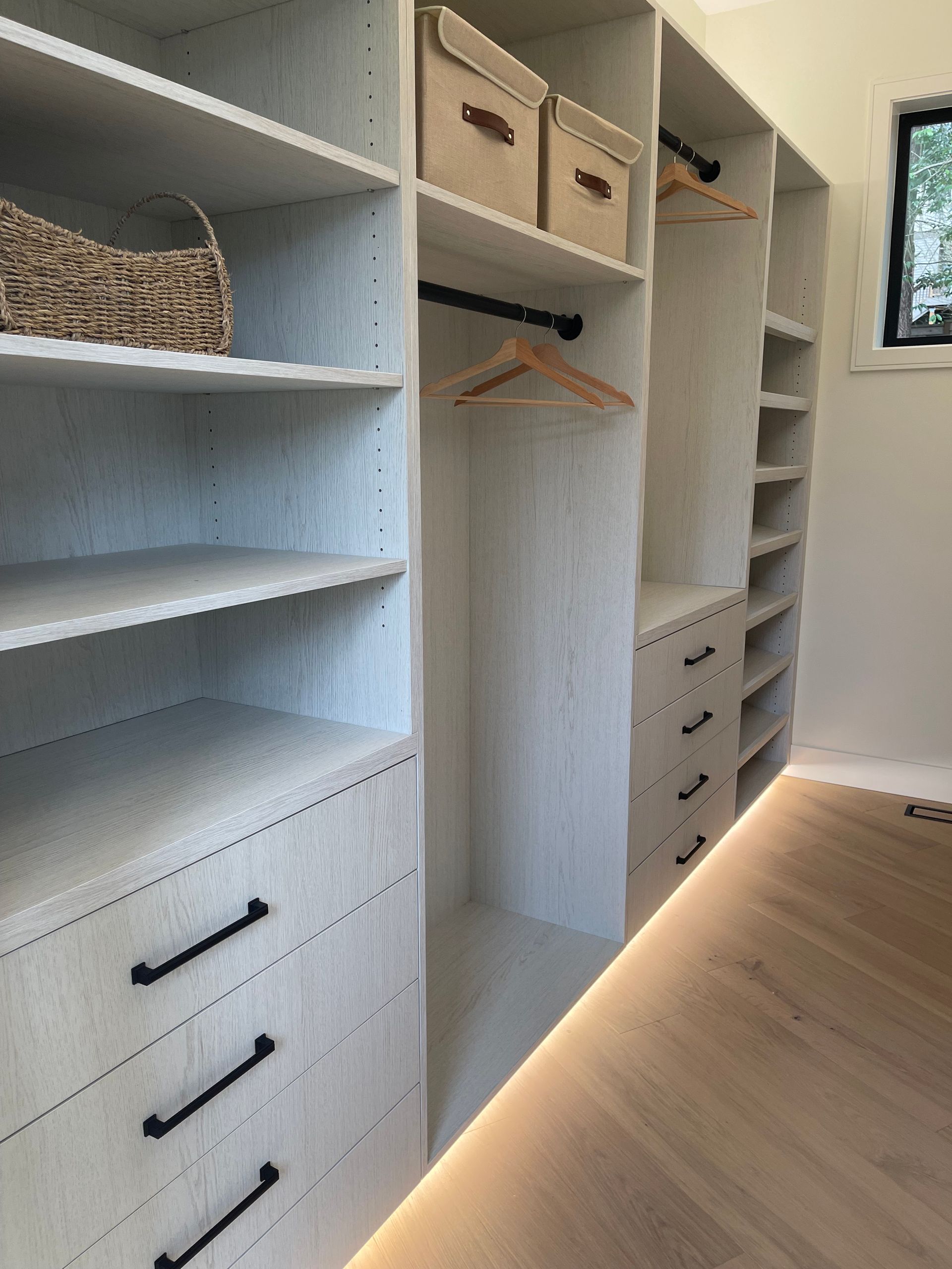 A walk-in closet featuring light wood-grain cabinetry, shelves, drawers with black handles, hanging rods, and floor lighting.