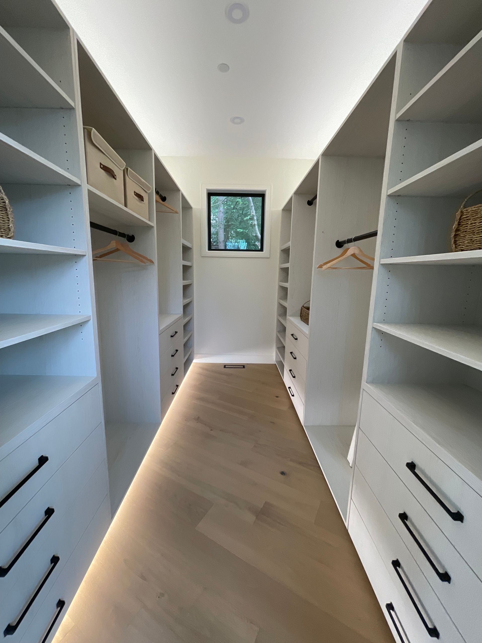 A walk-in closet with white shelving, drawer stacks, hanging rods, and light wood floors, lit by under-cabinet LED strips.