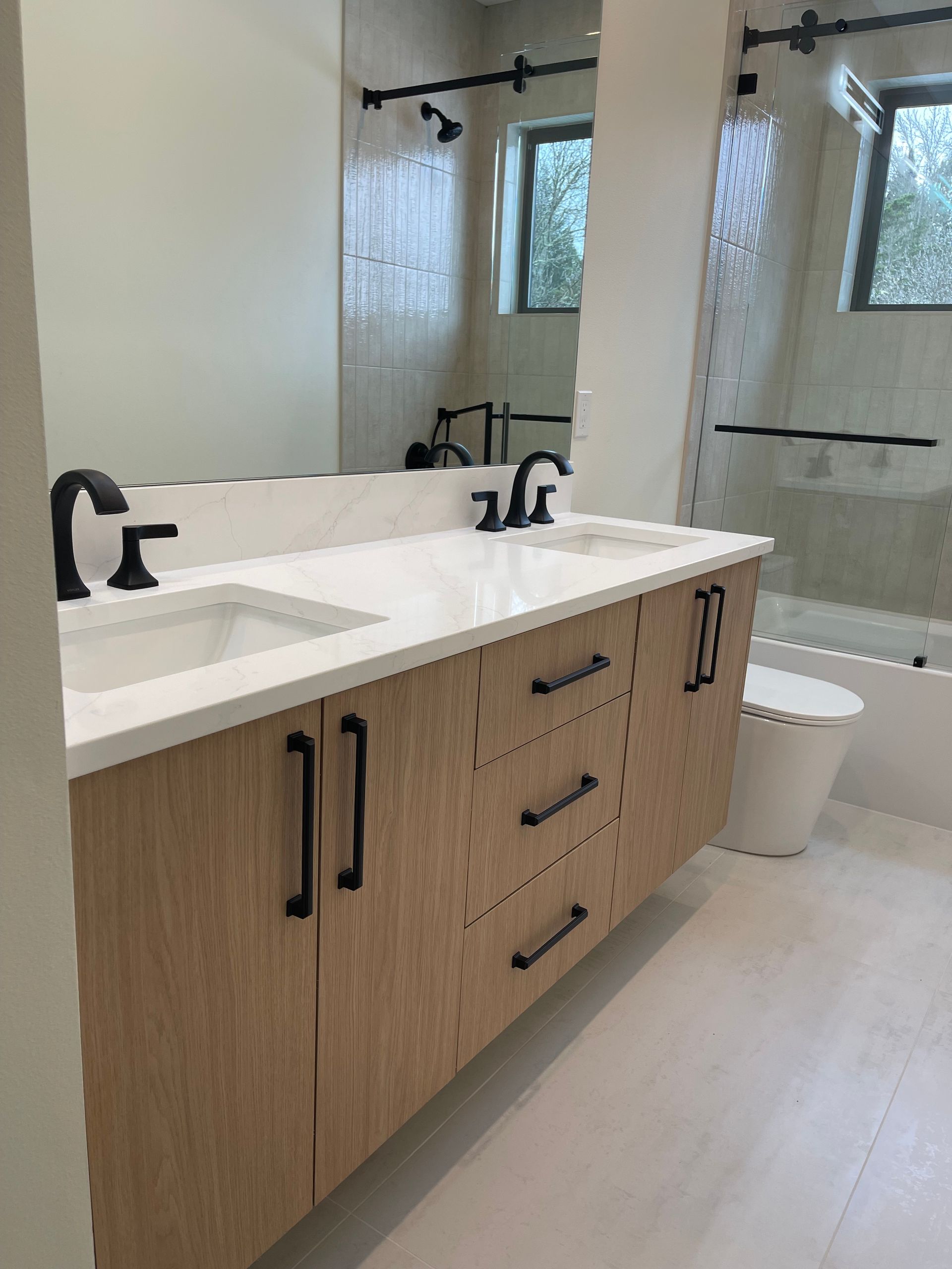 Modern double vanity with light wood cabinets, black hardware, white countertop, and rectangular sinks in a bathroom.