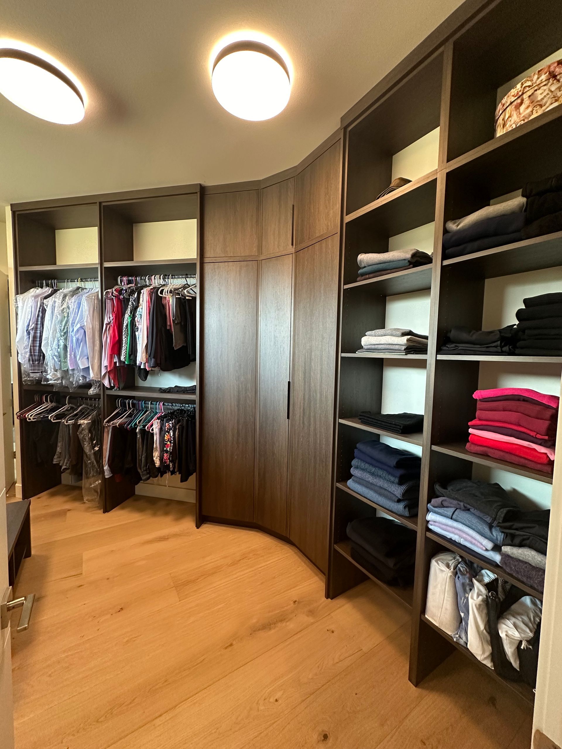 Walk-in closet with dark wood shelves and curved cabinets. Clothes hang on the left, folded clothes on the right.