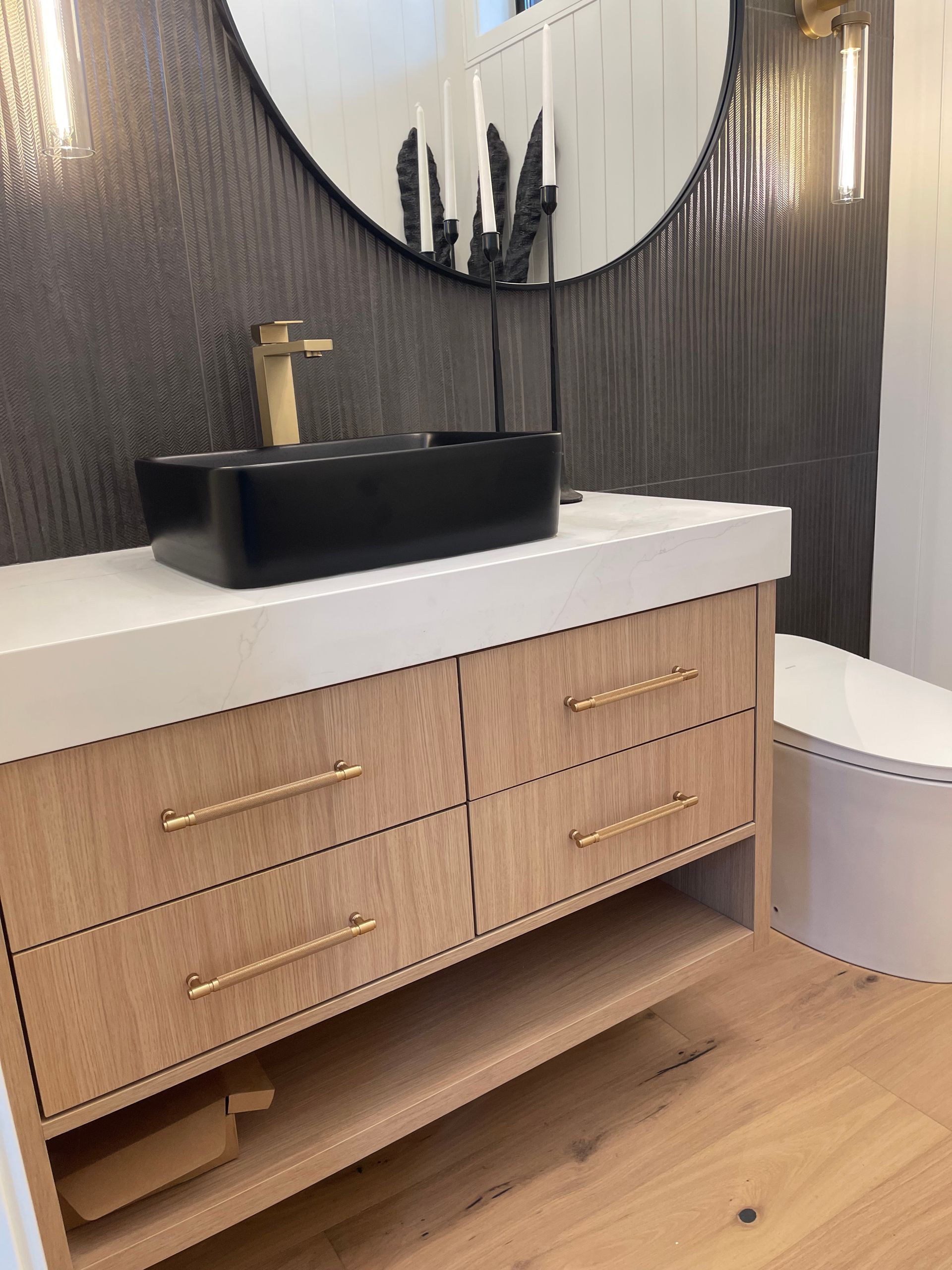 A modern bathroom vanity with a light wood finish, a black vessel sink, gold faucet, and a round mirror against a dark wall.