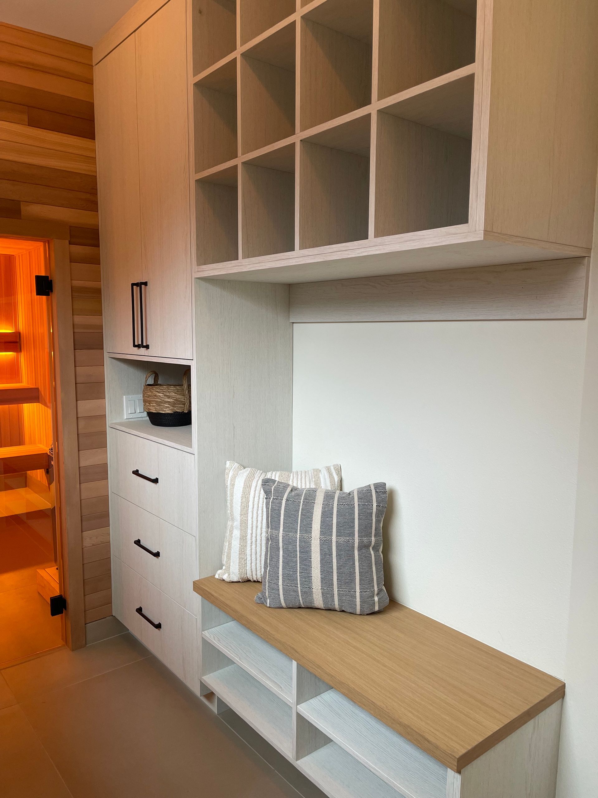 A mudroom storage unit featuring a white bench with a wood top, drawers, open cubbies, and a sauna visible to the left.