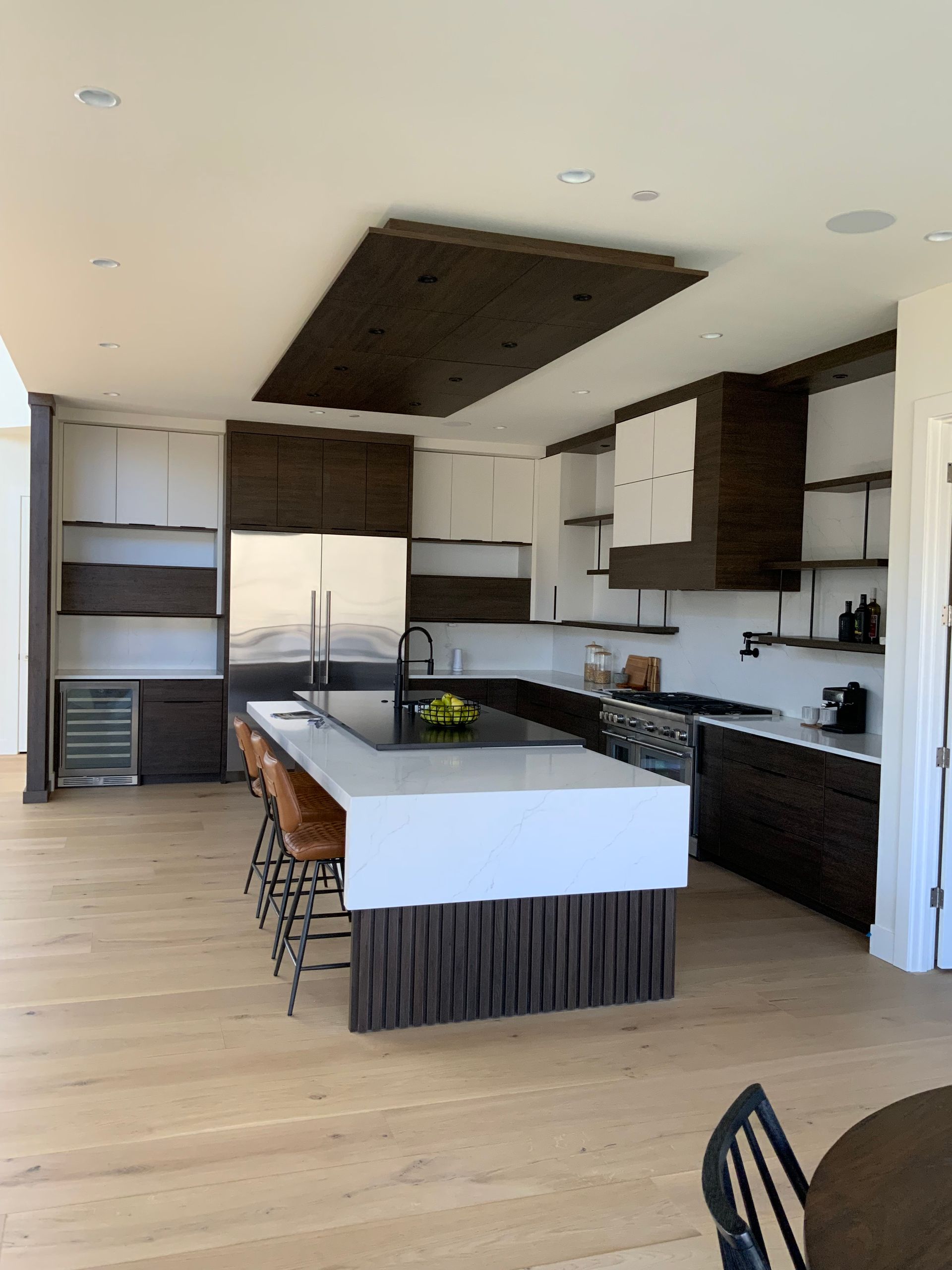 Modern kitchen with dark wood accents, white cabinets, island with stools, and light wood flooring.