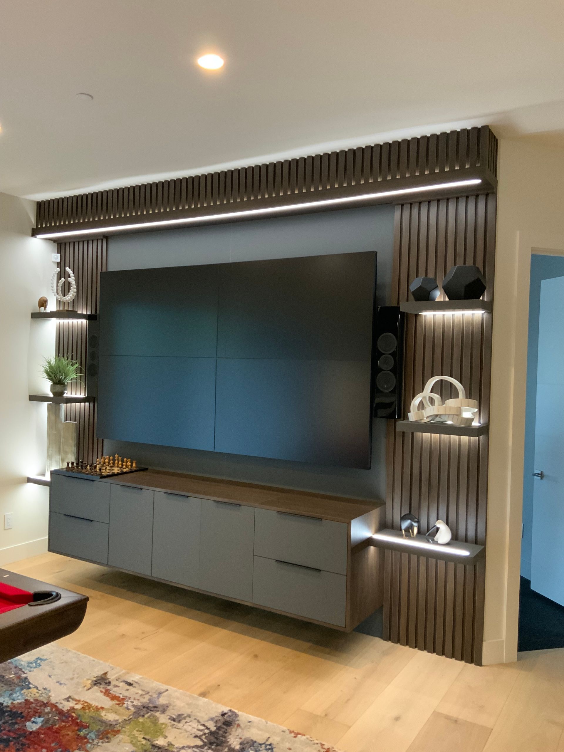 Modern media wall with large TV, floating shelves, and storage. Warm wood tones, LED lighting.