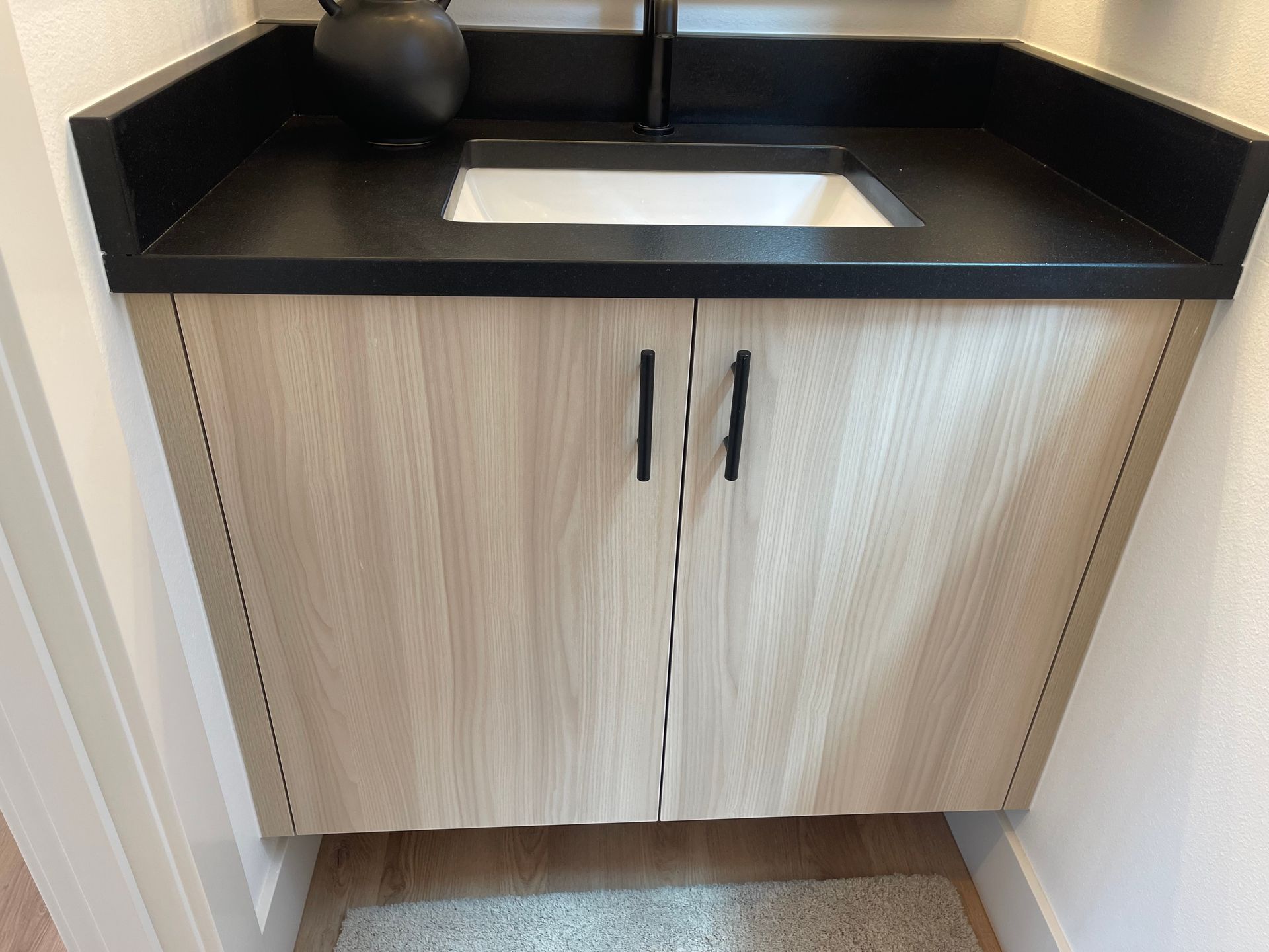 Bathroom vanity with light wood doors, black countertop, white sink, and black hardware.