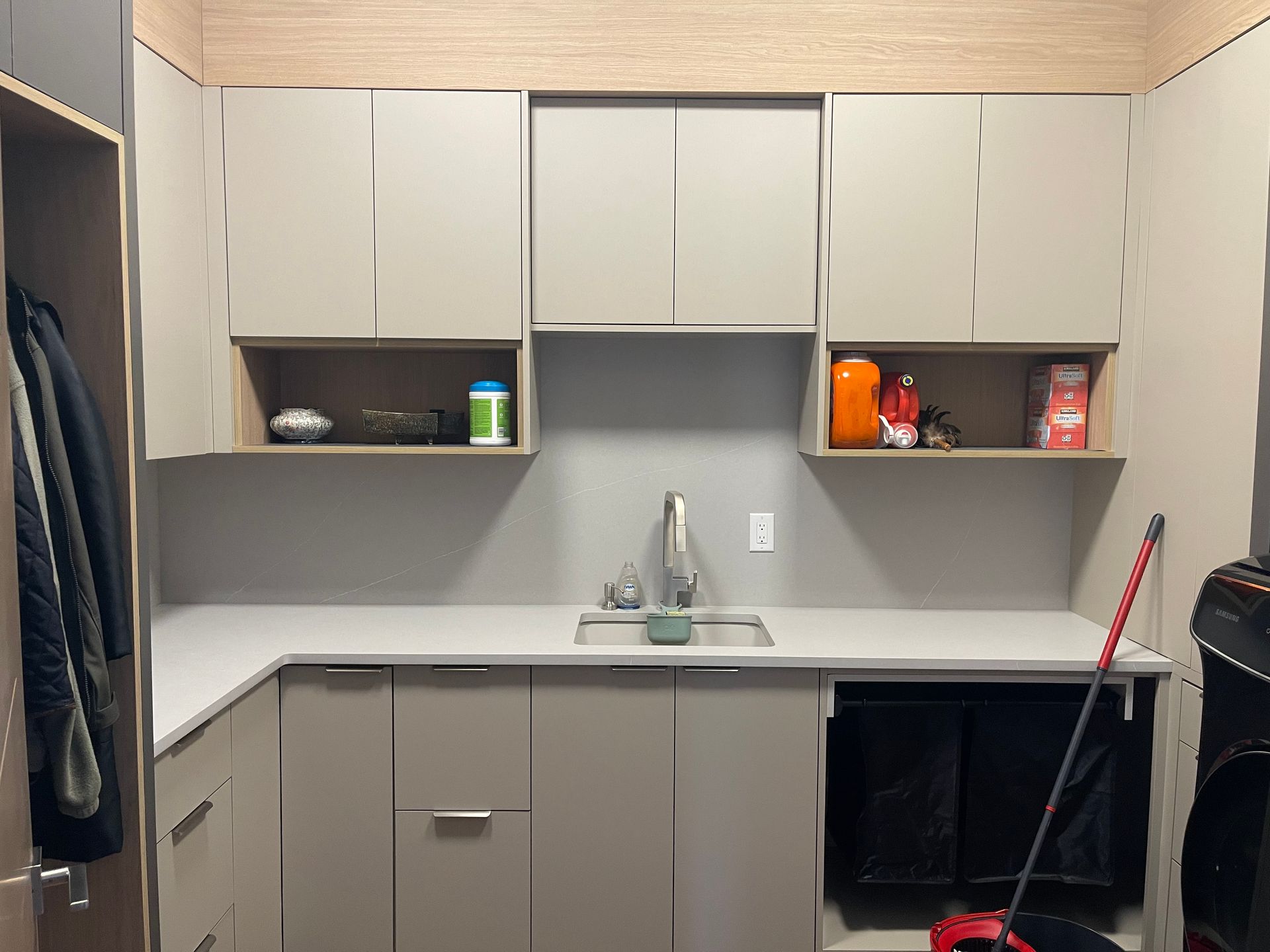 Laundry room with gray cabinets, sink, and countertop. Upper shelves hold cleaning supplies. Broom and coat on the side.