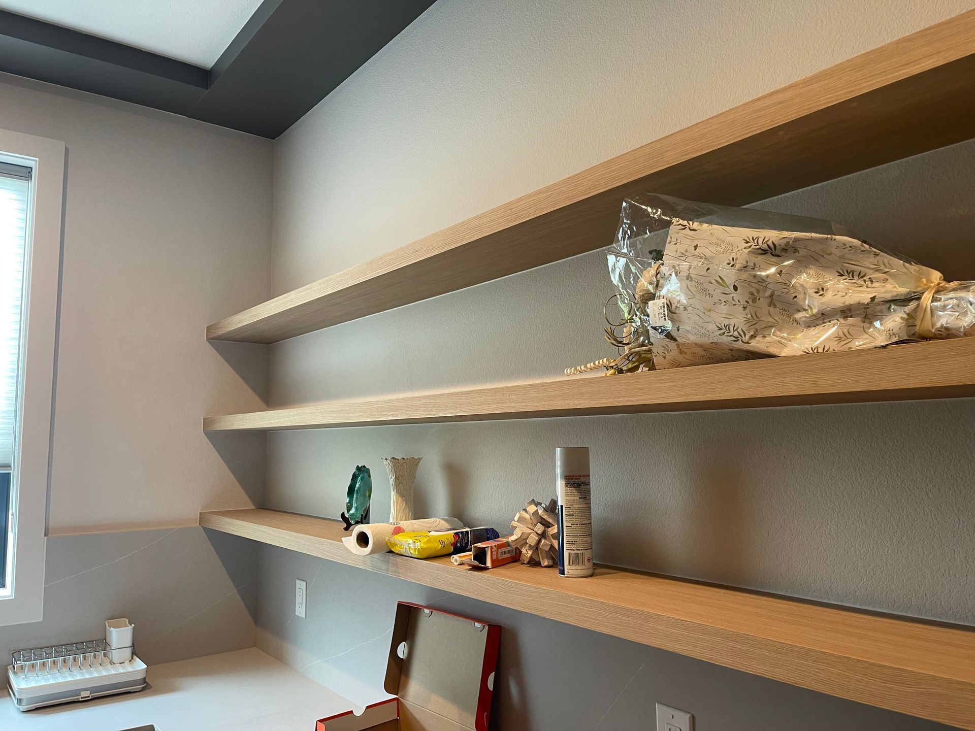 Three light-colored wooden shelves on a wall with items, including a bottle and toys, and a window.