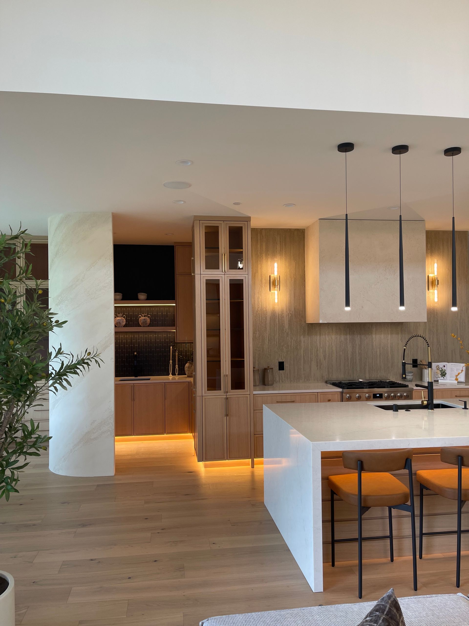 Modern open-plan kitchen with white island, wooden cabinets, warm lighting, and stools at the counter