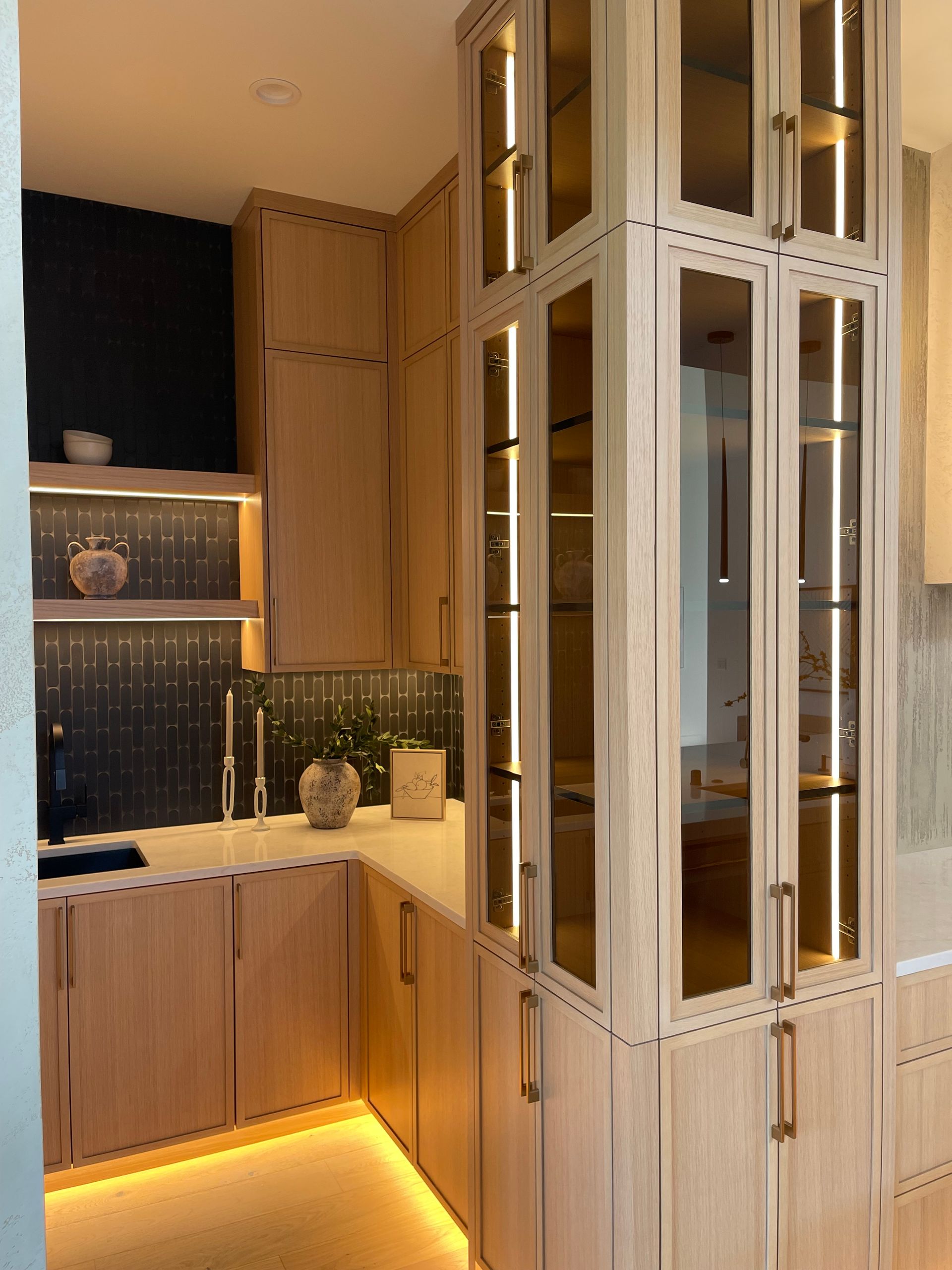 Modern kitchen with wood cabinets, black backsplash, and a tall glass-front pantry.