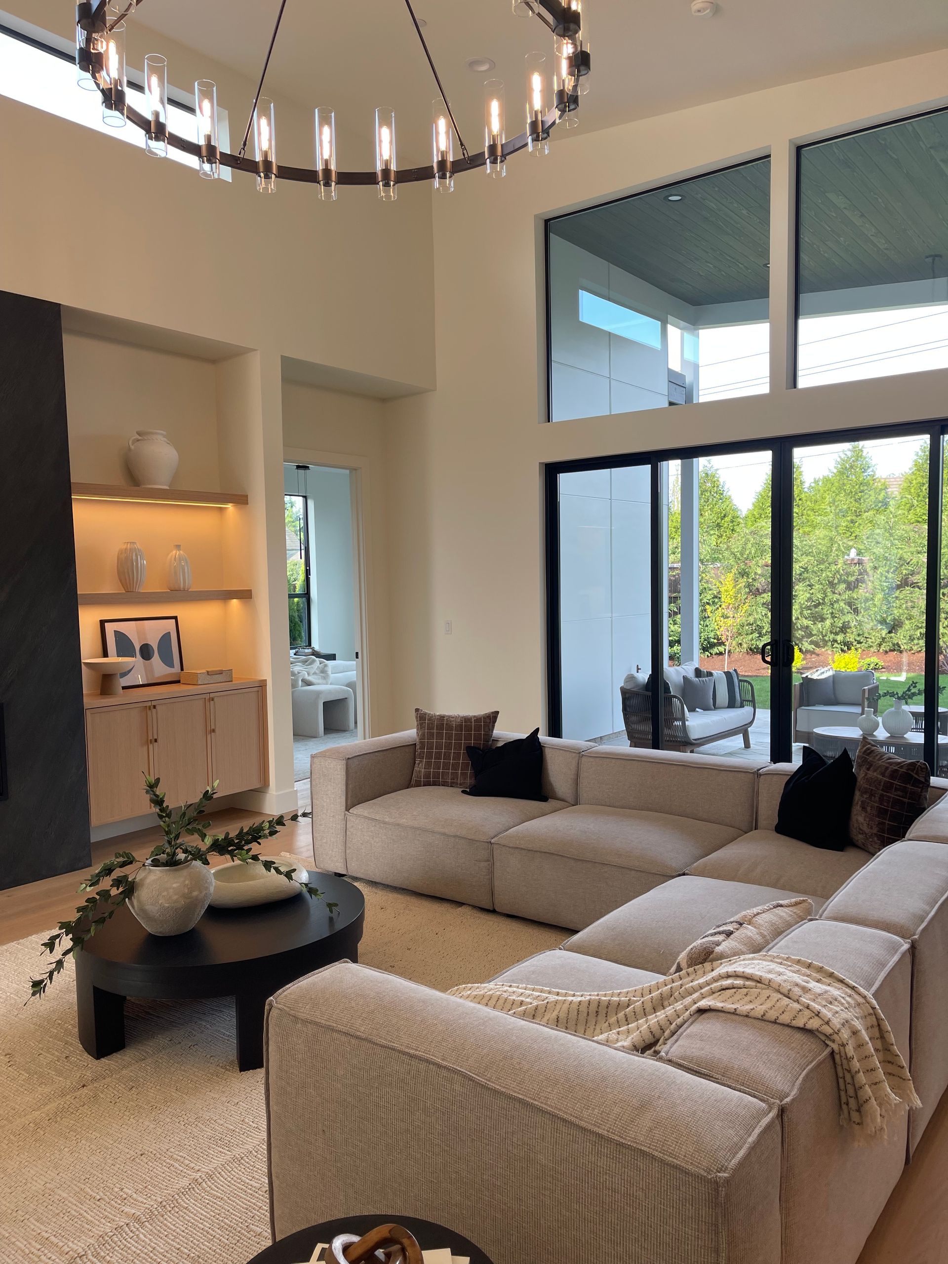 Modern beige living room with sectional sofa, chandelier, coffee table, and large windows overlooking trees