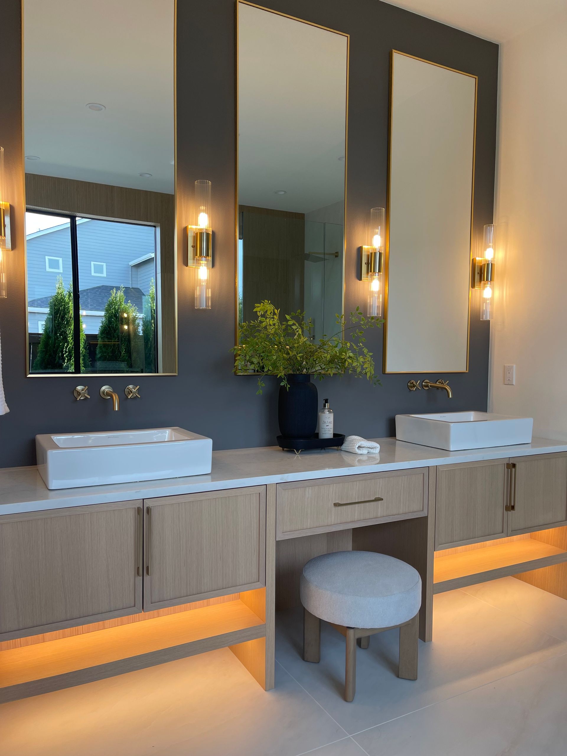 Modern restroom vanity with two sinks, large mirrors, and warm wall sconces above gray cabinetry.