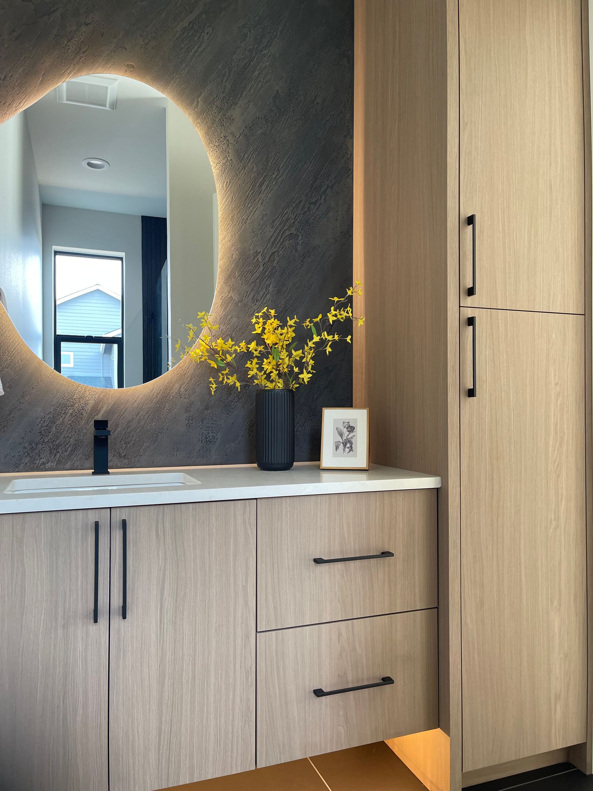 Modern entryway with light wood cabinets, round backlit mirror, and yellow flowers on a console.