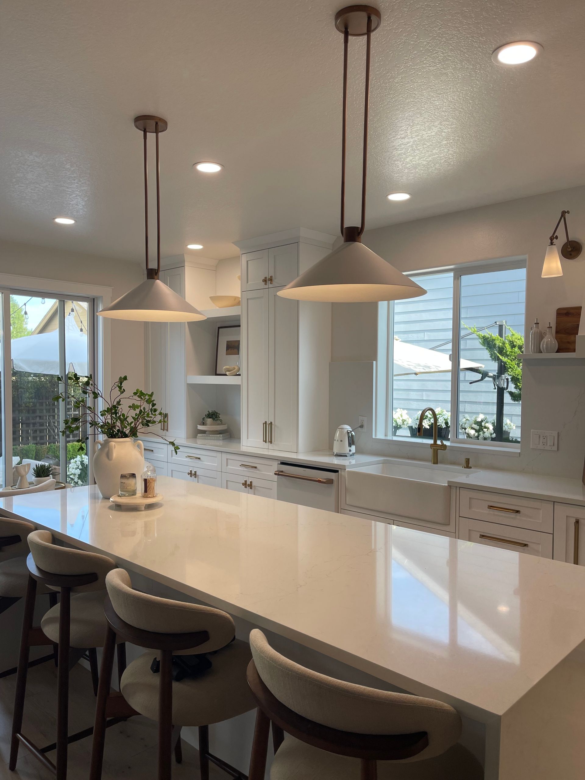 Bright modern kitchen with white island, pendant lights, bar stools, and large windows overlooking greenery