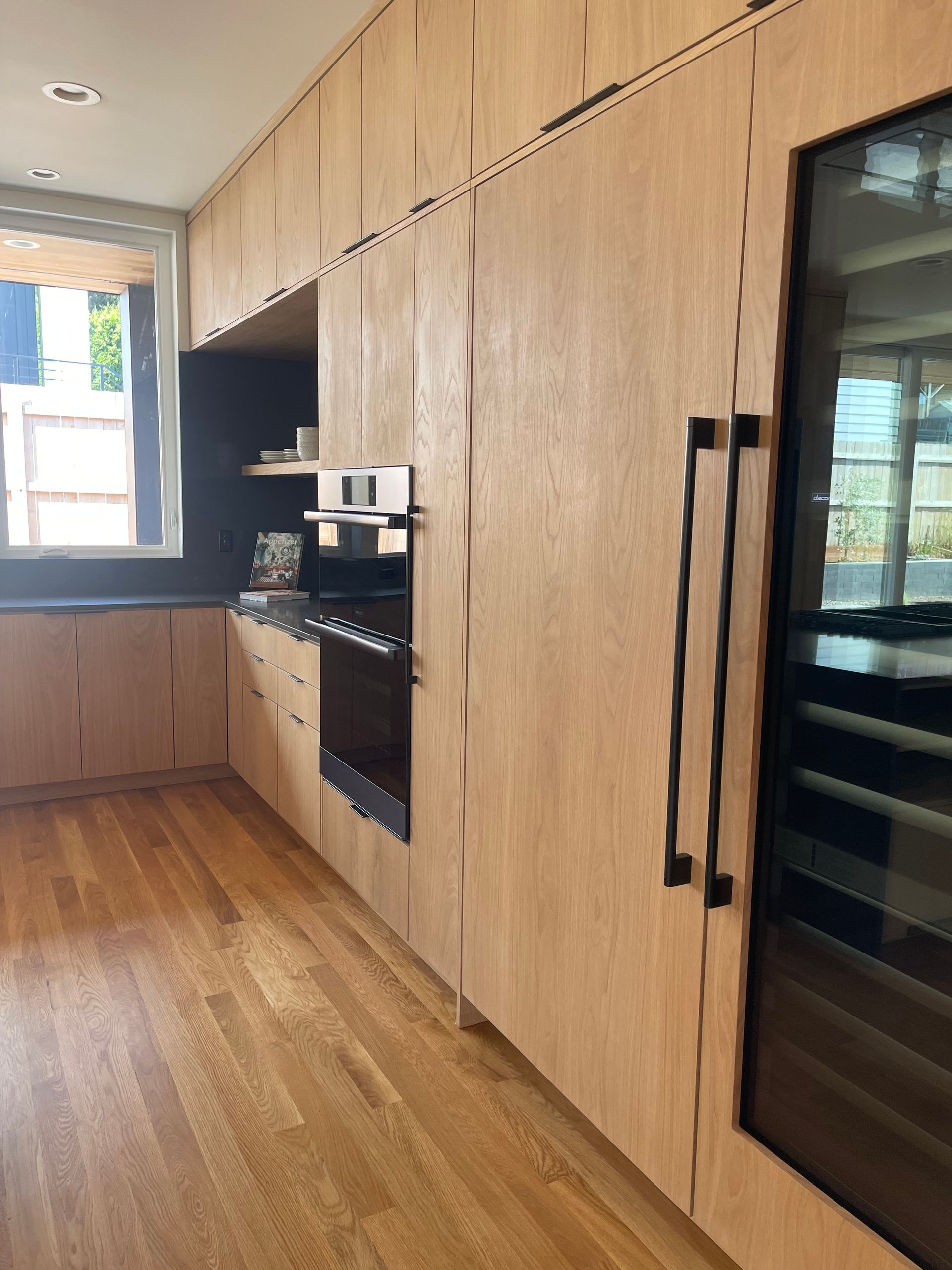 Wooden kitchen with light wood cabinets, built-in oven, and a large wine cooler with black handles.
